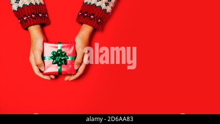 Female hands with a winter sweater holding a Christmas gift on a red background. Copy space Stock Photo