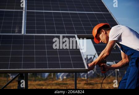 Using cordless screwdriver. Male worker in blue uniform outdoors with solar batteries at sunny day Stock Photo
