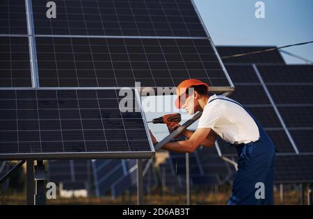 Using cordless screwdriver. Male worker in blue uniform outdoors with solar batteries at sunny day Stock Photo