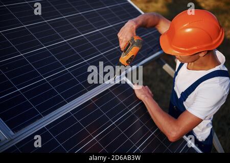 Using cordless screwdriver. Male worker in blue uniform outdoors with solar batteries at sunny day Stock Photo