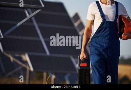 Holds case with equipment in hand. Male worker in blue uniform outdoors with solar batteries at sunny day Stock Photo