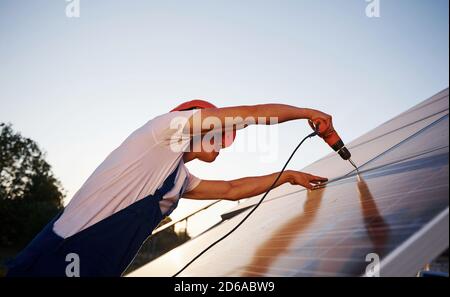 Using cordless screwdriver. Male worker in blue uniform outdoors with solar batteries at sunny day Stock Photo