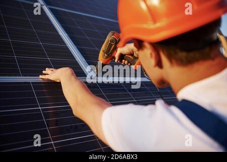 Using cordless screwdriver. Male worker in blue uniform outdoors with solar batteries at sunny day Stock Photo