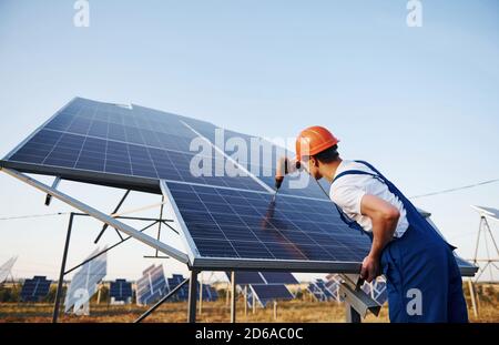 Using cordless screwdriver. Male worker in blue uniform outdoors with solar batteries at sunny day Stock Photo