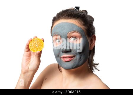 Cheerful caucasian woman with a clay or a mud mask on her face holding orange or tangerine over white background. Solving acne problems, cleansing, smoothing, rejuvenating the skin. The texture of a drying clay mask. Stock Photo