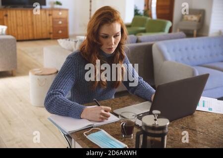 Woman taking notes while using laptop in the kitchen at home Stock Photo