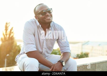 A dark-skinned guy in sunglasses sits in the park. Portrait of a late 20s black man sitting at the park on a summer day with filtered effect. Stock Photo