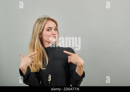 Pointing with hand at chest and staring. Shocked facial expression. Young attractive woman, dressed black sweater with green eyes, blonde hair, background Stock Photo