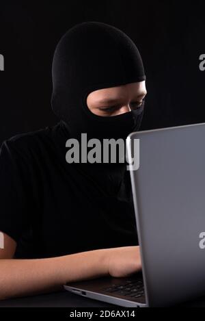Cyber terrorist in a mask in a mask on a black background behind a laptop Stock Photo