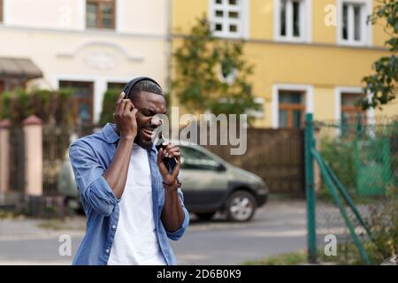 Walking in the street, relaxation, leisure. Young Afro-American man in headphones listening music on smart phone using music app. Portrait of smiling Stock Photo