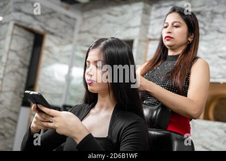 Female hairdresser standing and making hairstyle to cute lovely young woman in beauty salon, smiling young girl holding her mobile phone. Stock Photo