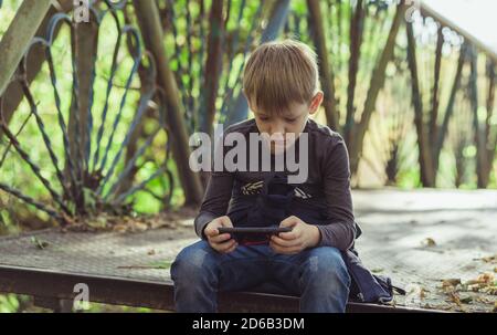 Focused boy using smartphone in park. Serious concentrated school aged boy in casual wear sitting on bench and playing game on mobile phone while Stock Photo
