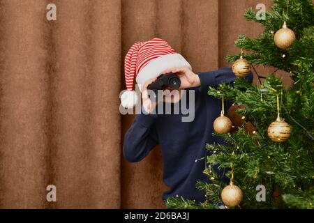 A boy peering out from behind a decorated Christmas tree, and looks through binoculars. On a brown curtain background. Stock Photo