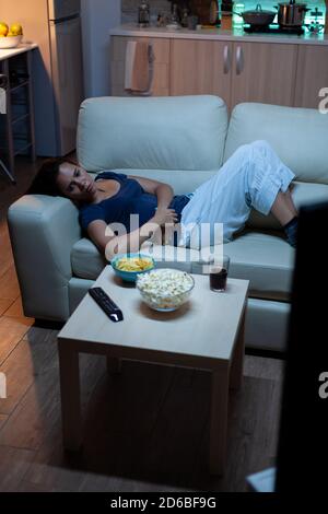 Young woman in pijamas resting lying on couch in front tv smiling. Happy, amused, lonely lady enjoying the evening sitting on comfortable sofa watching television eating popcorn and snacks Stock Photo