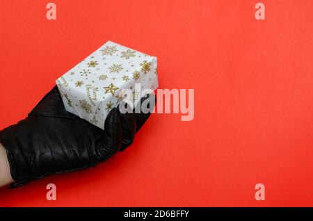 A hand in a black leather glove holds a wrapped Christmas present on a red background. Small rectangular box wrapped in white wrapping paper. Christma Stock Photo