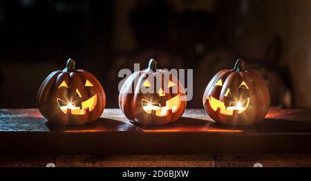 Three cheerful glowing Halloween pumpkins stand on a wooden table on a dark background. Stock Photo