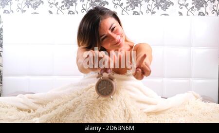 female rises in bed with alarm clock, enjoys the morning. The concept of starting a new day, relaxing on the weekend, no need to rush anywhere, good Stock Photo