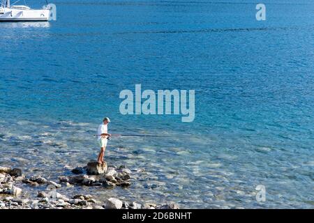 fisherman catches fish standing on a rock on the background of the sea. Fisherman standing on a rock sticking out of the blue sea Stock Photo