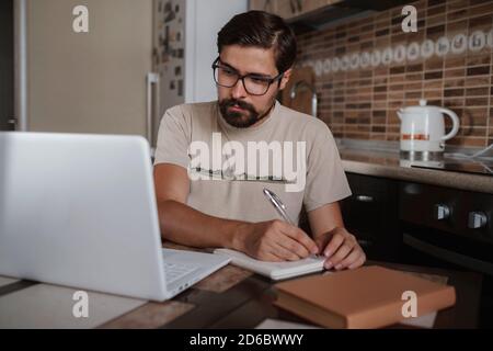 Concentrated millennial hipster guy wearing glasses, listening to favorite music while planning workday. Focused young biracial businessman watching e Stock Photo