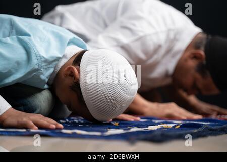 Muslim father and son praying or performing Salah while sitting on Prayer rug and touching head to mihrab or mosque printed on rug. Stock Photo