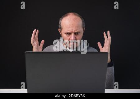 Man working from home office, angry face expression. Adult bearded sitting behind desk on black background, angry face, having business problem. Stock Photo