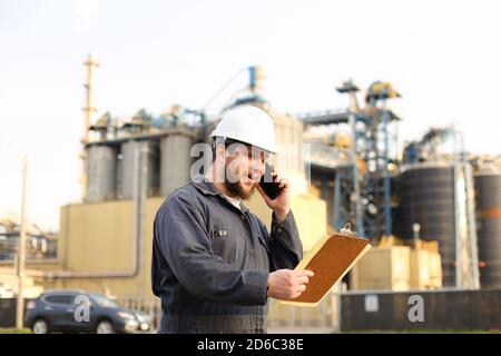 Industrial engineer talking by VHF walkie talkie and holding papers near factory outside. Stock Photo