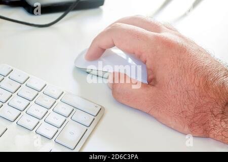 the white hand of a caucasian man working on a computer clicking a white mouse near a computer keyboard. Place of work and office. Wireless technology Stock Photo
