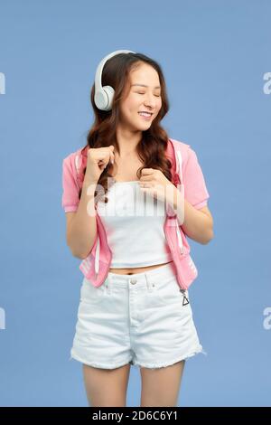 Energy Asian girl with headphones listening to music with closed eyes on blue background in studio. She wears white T-shirt, shorts. Stock Photo