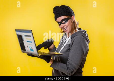 The concept of cybercrime and hacking. Portrait of a woman in a black hat, gloves and mask holding a laptop with a evil smile. Yellow background. Stock Photo