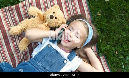 Pretty child girl laying on green lawn with her teddy bear talking on mobile phone. Stock Photo
