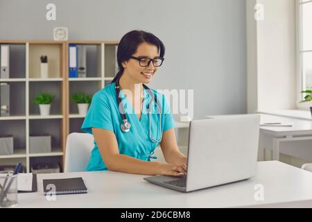 Young smiling doctor sitting and working on laptop online in medical clinic Stock Photo