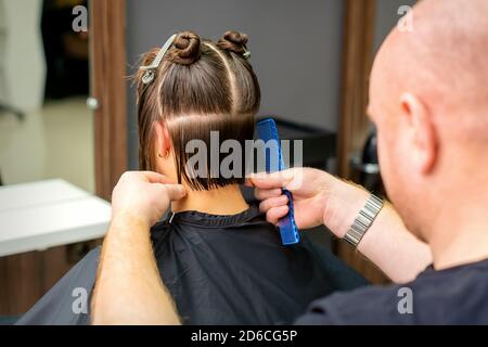 Male hairdresser cutting hair of young woman holding comb at hair salon. Rear view Stock Photo