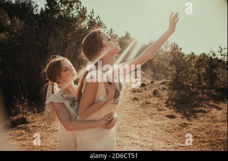 Two young beautiful girls in vintage identical dresses hug and sensually reach for the sun. Stock Photo