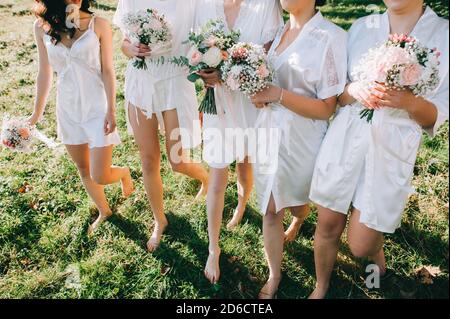 Faceless bridesmaids and bride in satin robes with bouquets in hands walk barefoot on the grass. Stock Photo