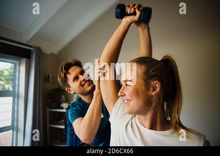 Male physiotherapist correcting arm alignment of female patient holding weights sitting in pilates studio. Stock Photo