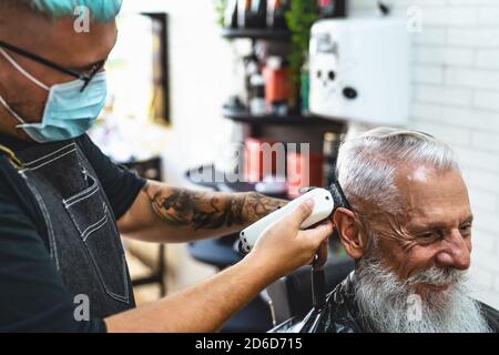 Male hairdresser cutting hair to hipster senior client while wearing face surgical mask - Young hairstylist working in barbershop during corona virus Stock Photo