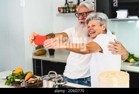 Happy senior couple taking selfie while cooking together at home - Elderly people having fun preparing health lunch in kitchen Stock Photo