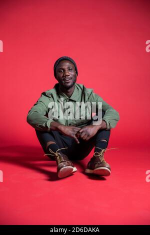 Relaxed young black man sitting on floor during studio photoshoot. Isolated. Full length. Vertical. Stock Photo