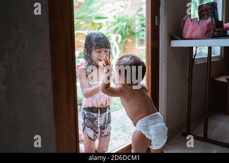 Little sister stood looking at her older sister outside through the glass door while playing together Stock Photo