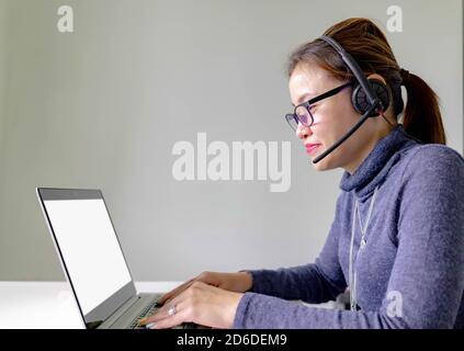 Happy smiling young asian support phone operator in headset with laptop, on grey background with laptop showing white screen Stock Photo