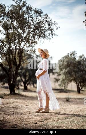 Pregnant woman wearing hat and white dress among cork trees Stock Photo