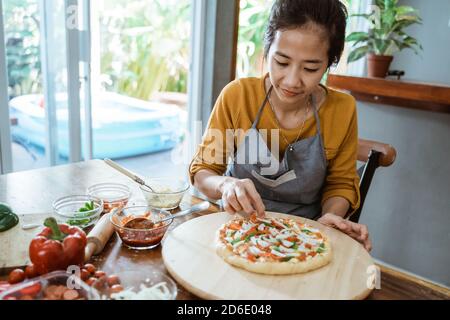 Happy attractive young adult woman lady housewife baker wear apron making homemade pizza on kitchen table at home Stock Photo