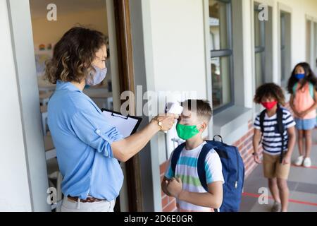 Female teacher wearing a face mask measuring temperature of a boy Stock Photo