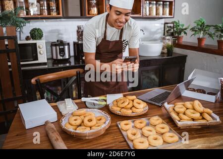 male chef smiles while taking pictures of donuts using a smartphone for online marketing of homemade products Stock Photo