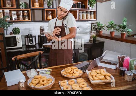Asian Chef calls using a smartphone while matching notes on a donut order from a notebook with a laptop, working on making homemade donuts Stock Photo