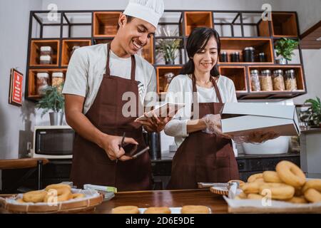 male and female chefs smile as they take notes, prepare donut orders, and pack donuts, marketing homemade donuts Stock Photo