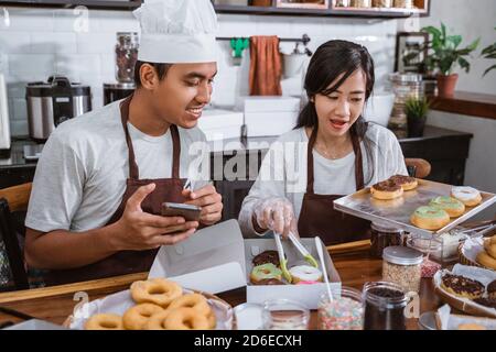 A male chef wearing an apron using a cellphone and a female chef preparing a donut order, placing it in a packing box, after making donuts together in Stock Photo