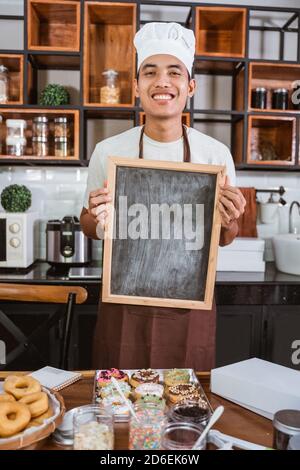 male chef wearing a smiling apron holds a blackboard while standing in the kitchen room Stock Photo