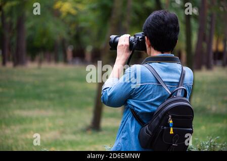 Woman photographer taking pictures outdoors. Young brunette with short hair dressed in blue denim shirt with backpack holding camera. Rear view Stock Photo