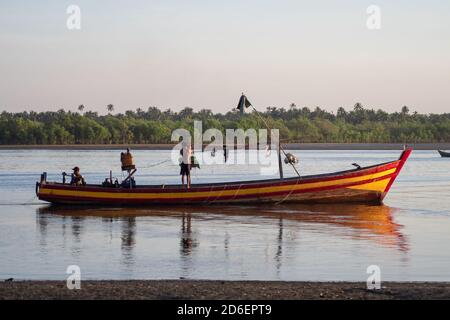 Chaung Thar, Myanmar - December 26, 2019: A traditional wooden boat manned by two local people by the shore of a river lit up by the sunset Stock Photo